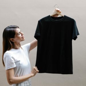 A cheerful woman in a white t-shirt holding a black t-shirt on a hanger against a gray background.